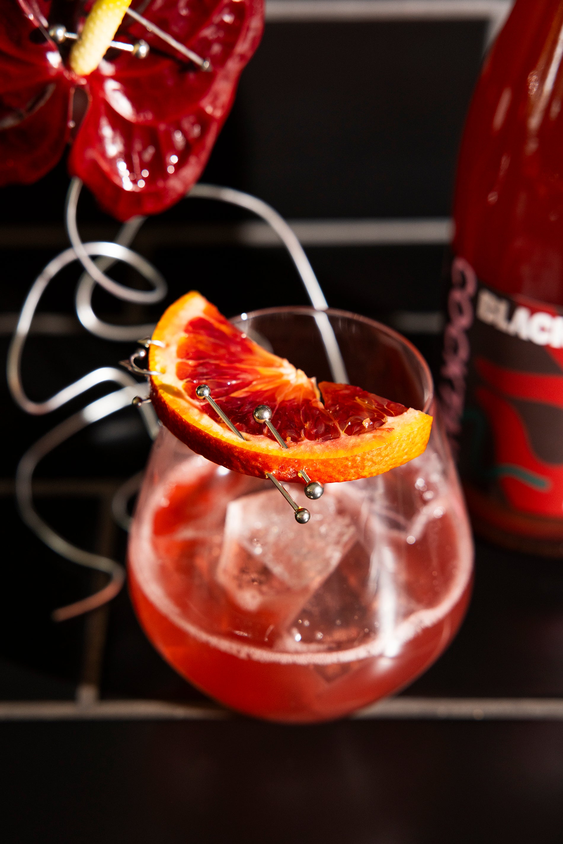 Idle Hands Drinks Co Black Cherry French Martini cocktail with blood orange slice and ice cubes in a glass, garnished with a red flower, against a dark background.